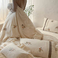 A person wearing a beige ribbed sweater is shown from the waist down, holding up a cream-colored bedding set with delicate floral embroidery and striped details. The set includes a duvet cover and matching pillowcases, styled on a bed in a softly lit, rustic bedroom.
