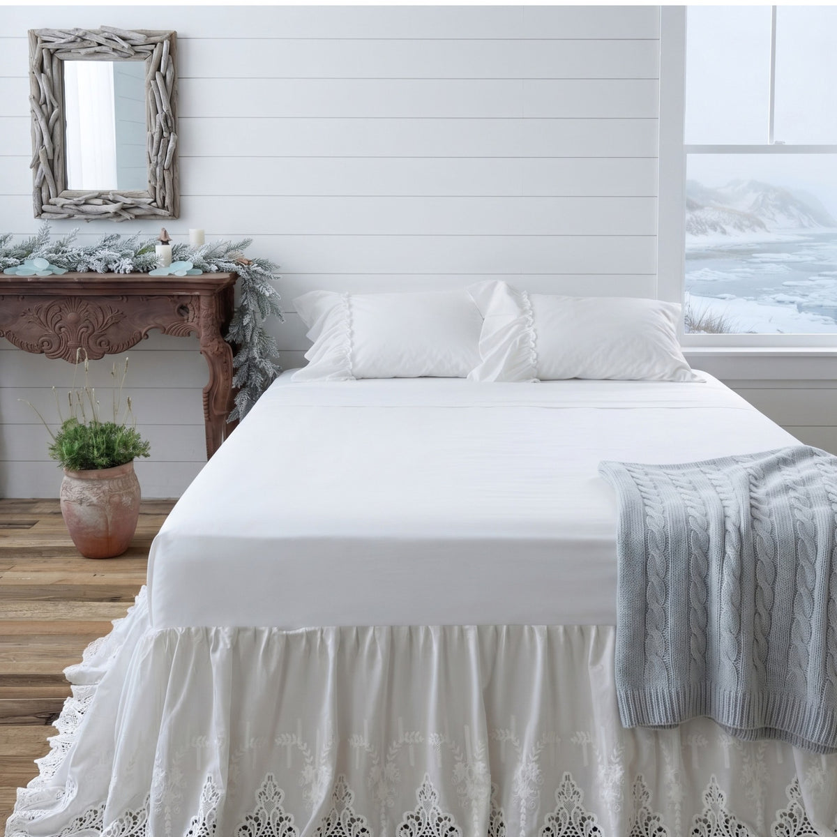 A neatly made bed dressed in white bedding and a white ruffled bed skirt with lace trim, styled in a bright bedroom with a wooden console table, mirror, and window view.