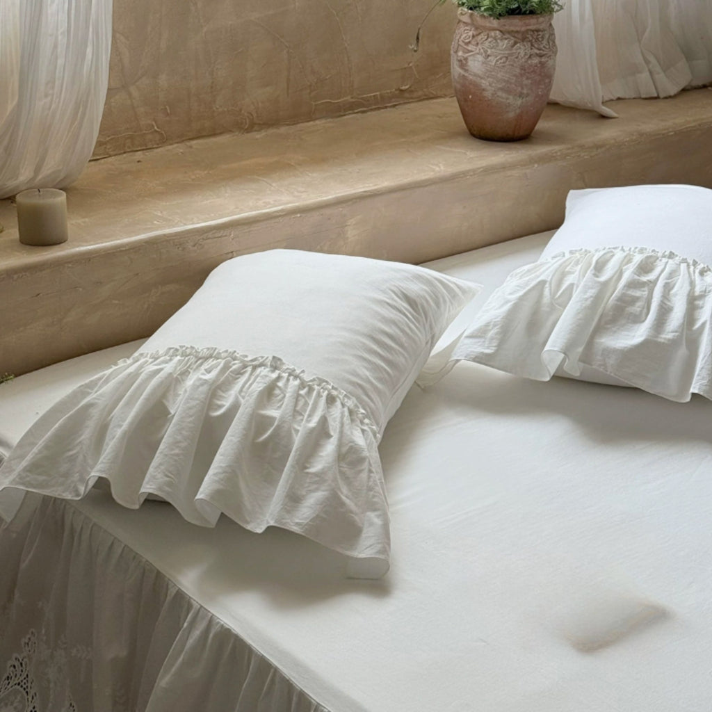 White ruffled pillowcases on a bed with a matching white bed skirt, styled in a warm, rustic bedroom setting with a stone ledge and a potted plant.