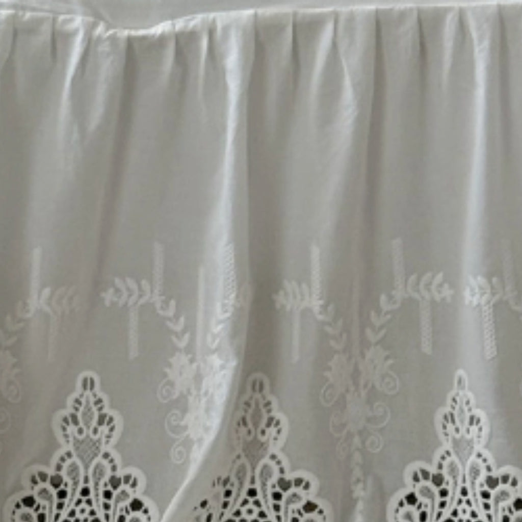 White pleated fabric with embroidered floral and leaf patterns and a scalloped lace border at the bottom.