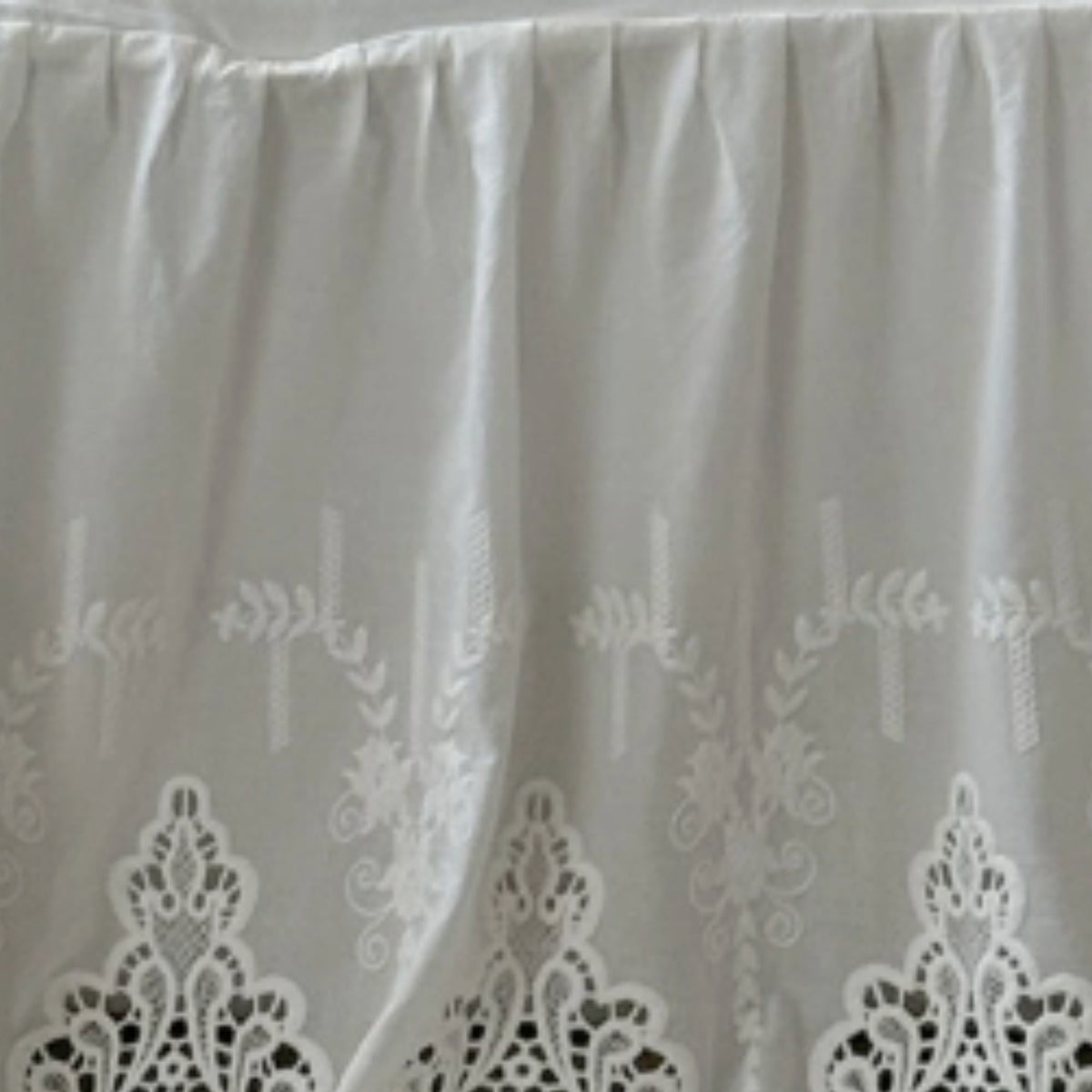 White pleated fabric with embroidered floral and leaf patterns and a scalloped lace border at the bottom.