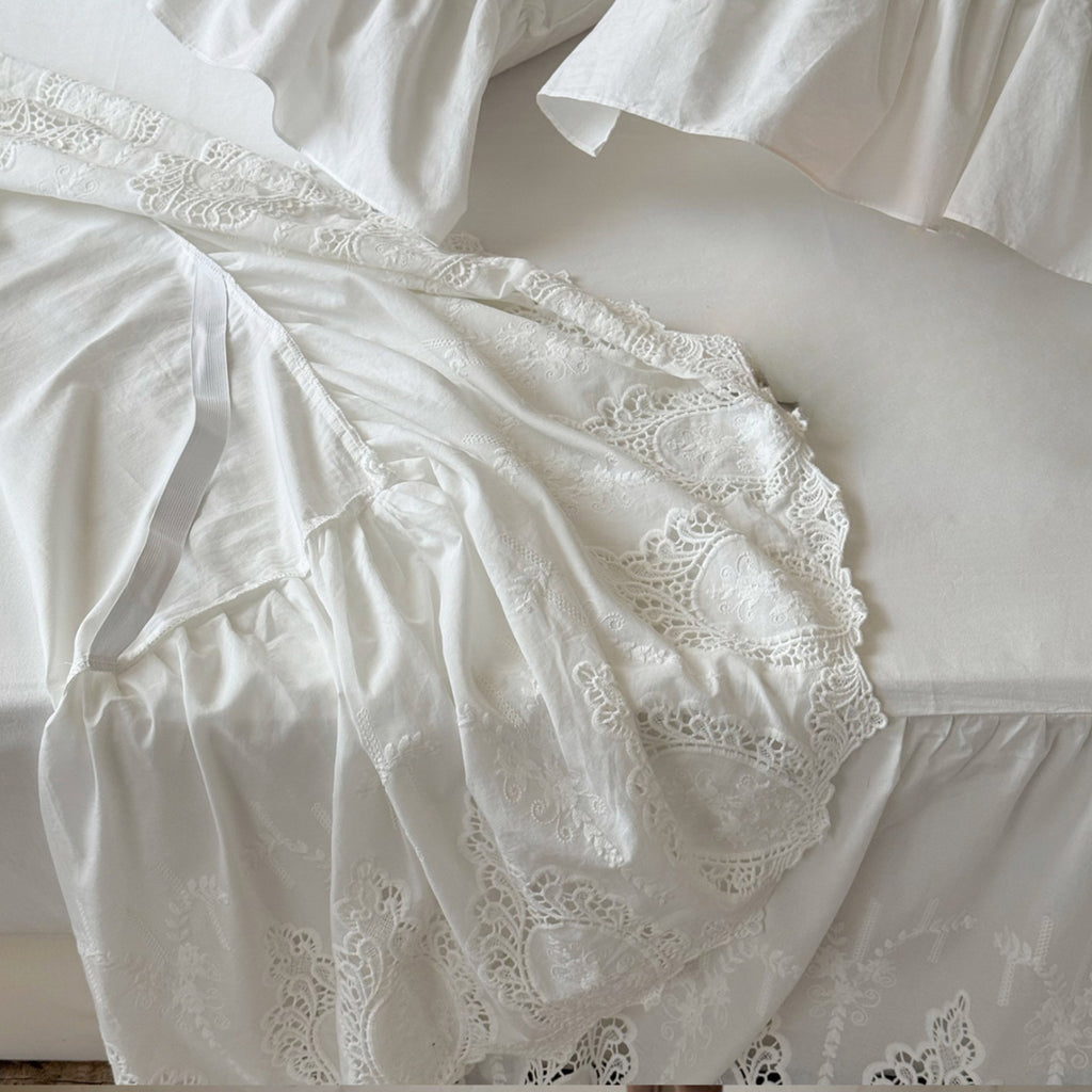 White lace-trimmed bedding set draped on a bed, including a sheet, pillowcases, and a matching skirt with delicate floral lace borders.