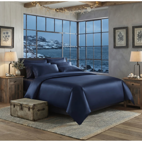 A navy blue silk bedding set, including a comforter and matching pillows, styled on a bed in a rustic bedroom with wooden nightstands, lamps, and a large window showing an ocean view.