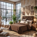 A cozy bedroom scene with a bed dressed in a brown quilted blanket and matching pillows, set in a warm, rustic room with large windows, plants, wall art, and a macrame hanging.