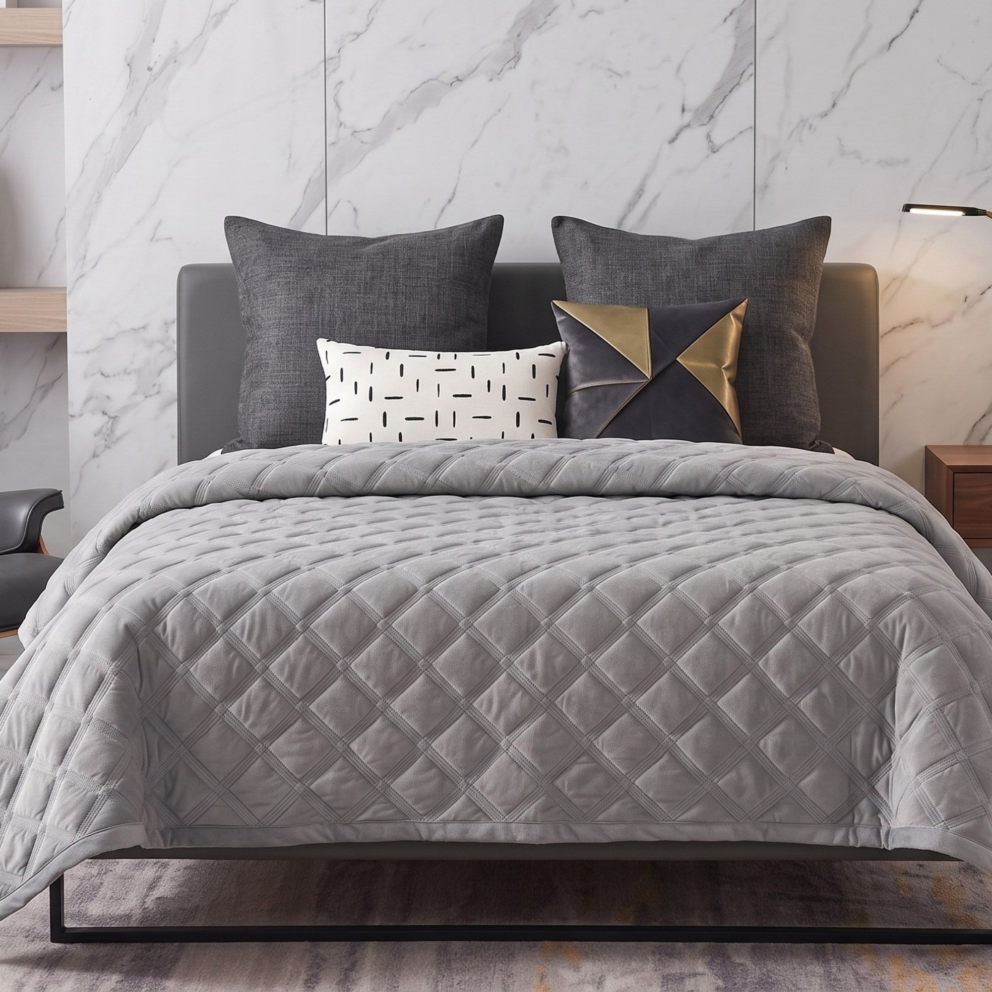 A gray quilted comforter on a bed with dark gray pillows, a white accent pillow with black markings, and a geometric gold and navy throw pillow, set against a white marble wall.