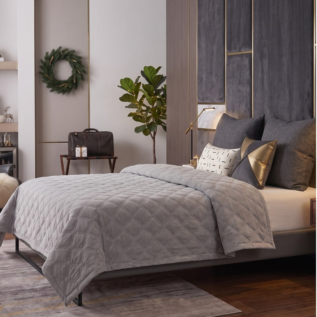 A styled bedroom scene showing a bed made up with a light gray quilted comforter, white sheets, and several pillows including dark gray, metallic gold, and a small white patterned accent pillow. The room includes a dark wood floor, a gray upholstered headboard, a side table with a lamp and bag, a plant, and a green wreath on the wall.