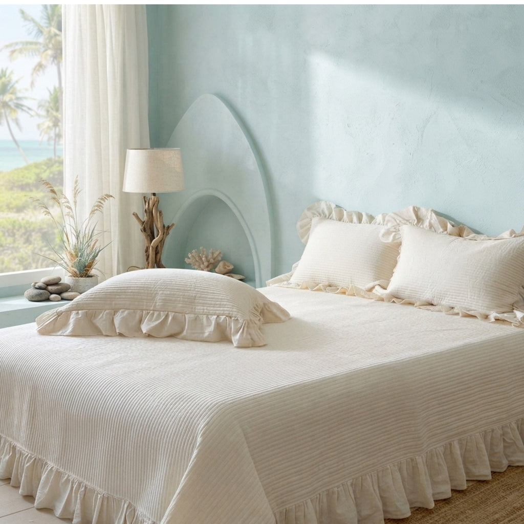 A bed dressed in cream-colored ruffled bedding, including a textured coverlet and matching pillows, styled in a coastal-themed bedroom with a light blue wall, a window with palm trees, and a bedside lamp.