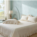A bed dressed in cream-colored ruffled bedding, including a textured coverlet and matching pillows, styled in a coastal-themed bedroom with a light blue wall, a window with palm trees, and a bedside lamp.