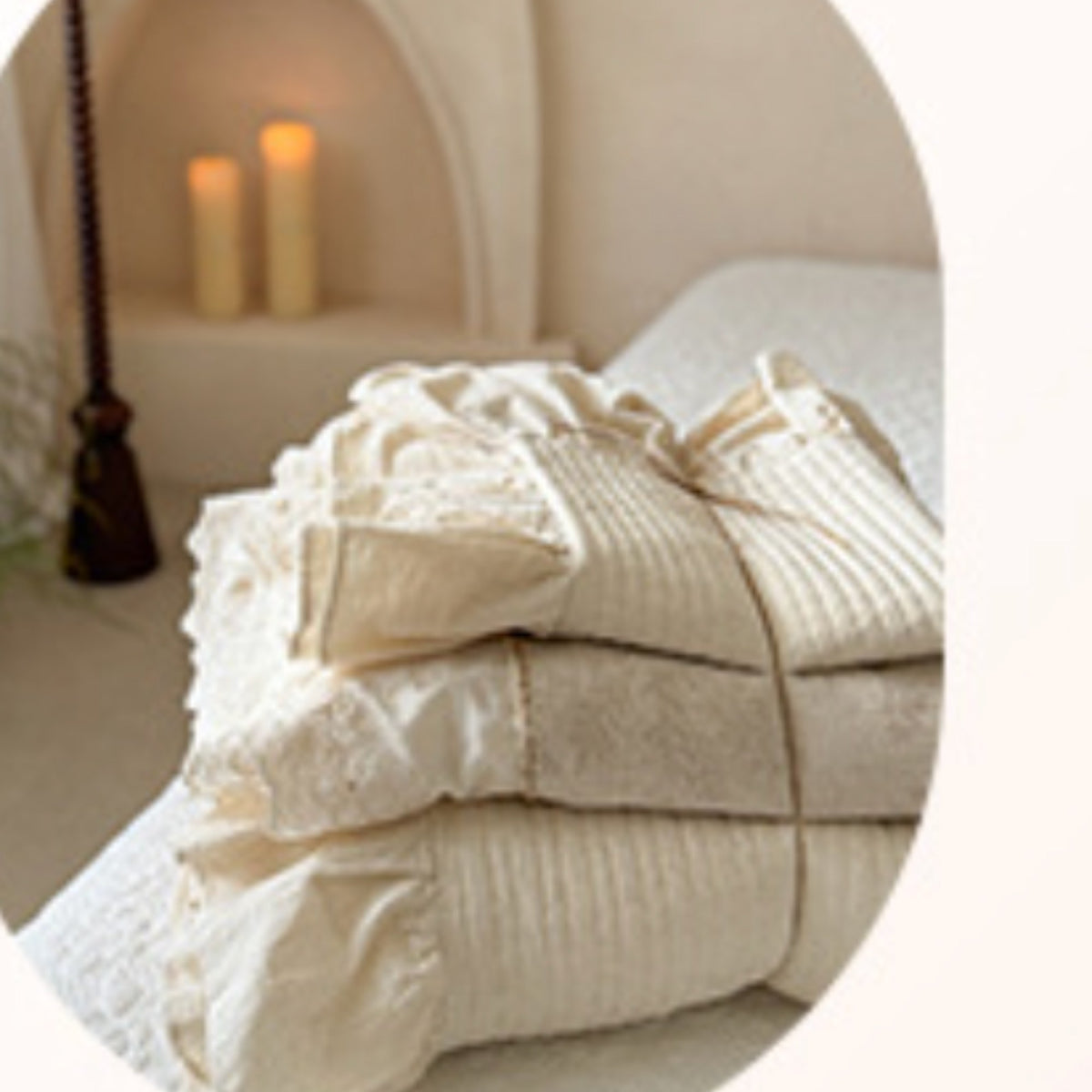 A stack of cream-colored bedding, including a ruffled pillowcase and textured sheets, styled on a bed in a cozy bedroom setting with warm candlelight.