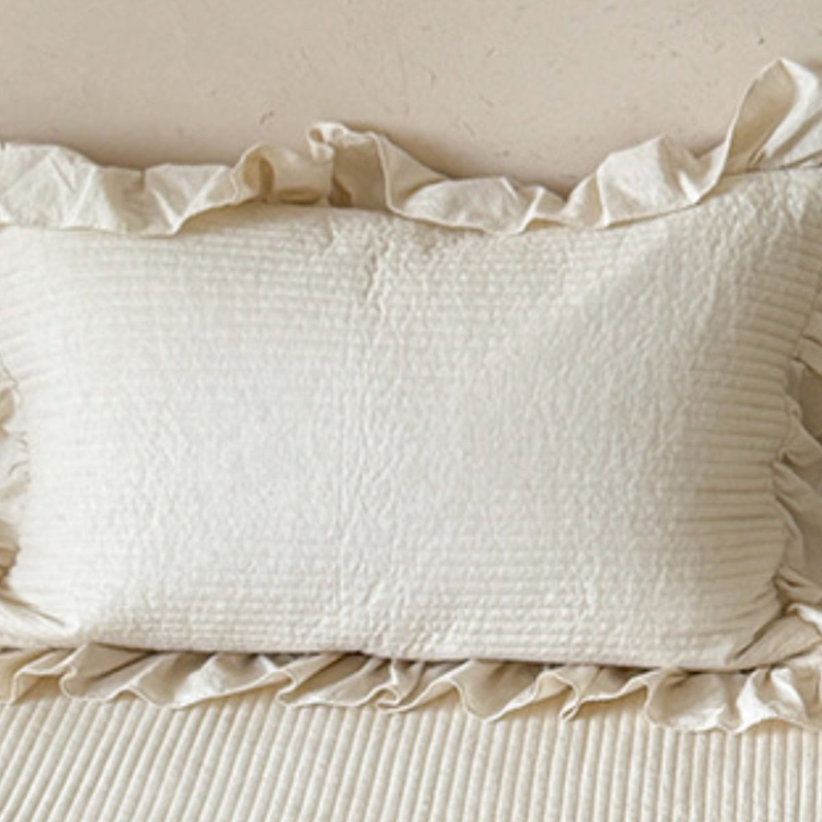 An off-white textured pillowcase with ruffled trim, resting on a matching ribbed bedspread against a neutral beige wall.
