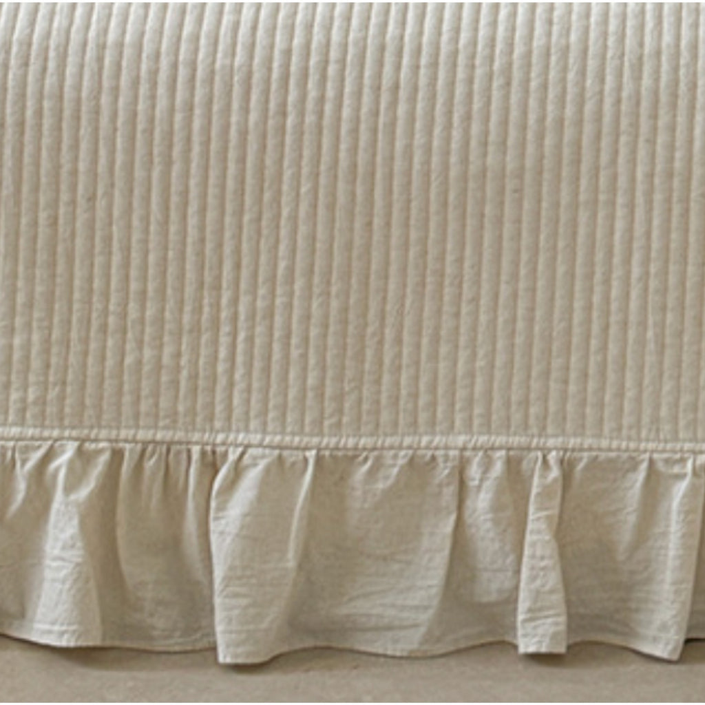 A close-up of a cream-colored bed skirt with vertical channel tufting and a ruffled hem, shown against a neutral floor background.