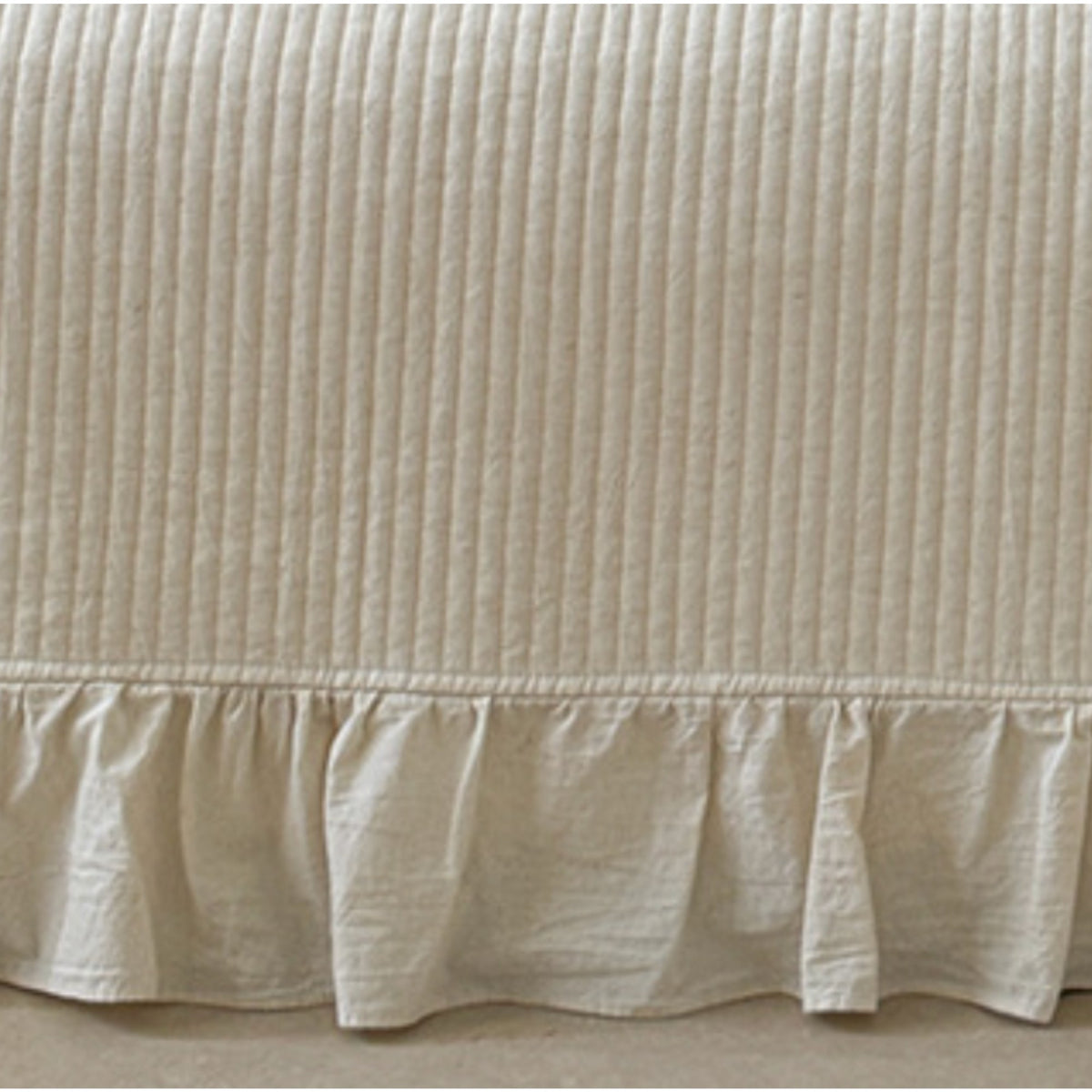 A close-up of a cream-colored bed skirt with vertical channel tufting and a ruffled hem, shown against a neutral floor background.
