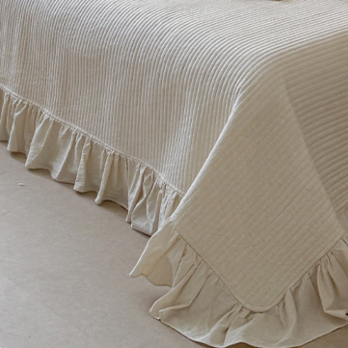 A cream-colored bedspread with a ruffled skirt and subtle vertical textured stripes, draped over a bed in a softly lit room.