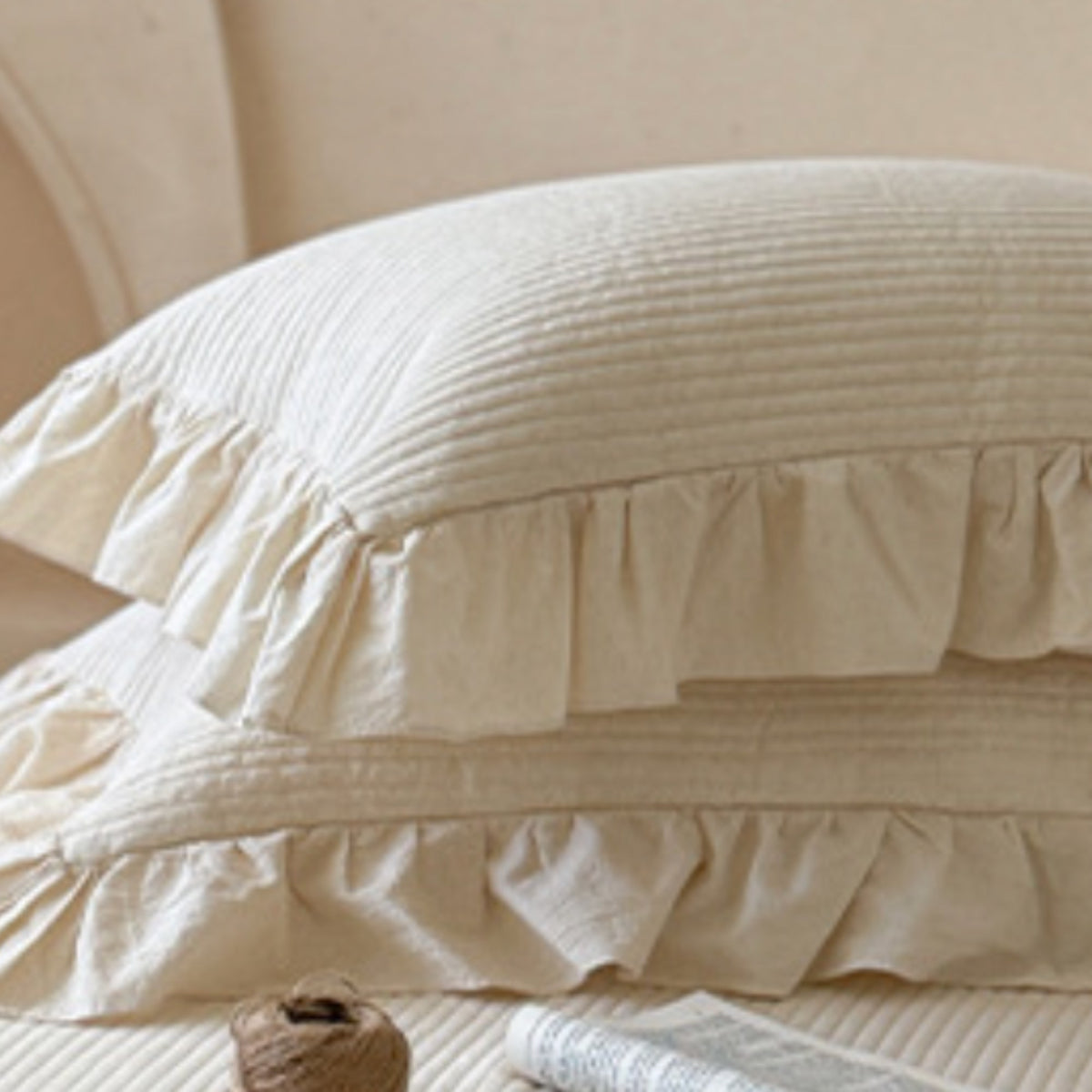 Two cream-colored ruffled pillowcases stacked on a matching bedspread, with a soft neutral background and a small decorative spool in the foreground.