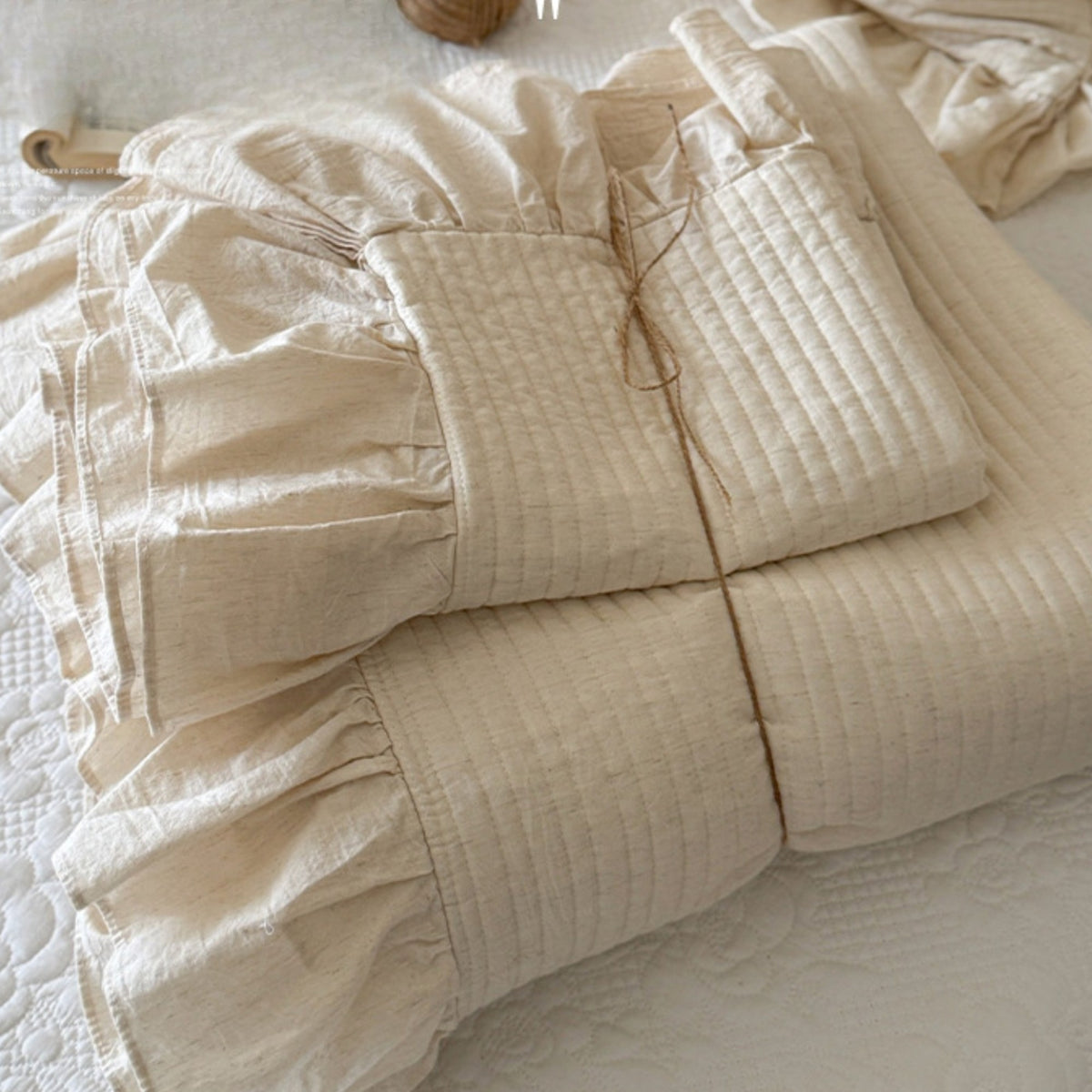 A folded cream-colored quilted bedding set with ruffled details, displayed on a white textured quilted bedspread.