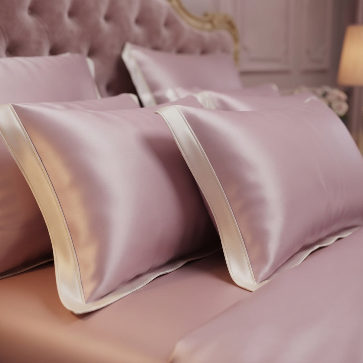A close-up of a bed dressed in matching silk pillowcases and sheets in a soft dusty pink color with contrasting cream silk piping, set against a tufted pink headboard in a softly lit bedroom.