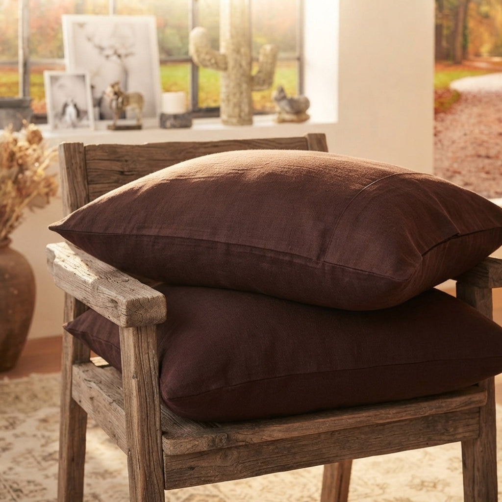Two dark brown linen-textured throw pillows stacked on a rustic wooden chair in a warm, sunlit room.