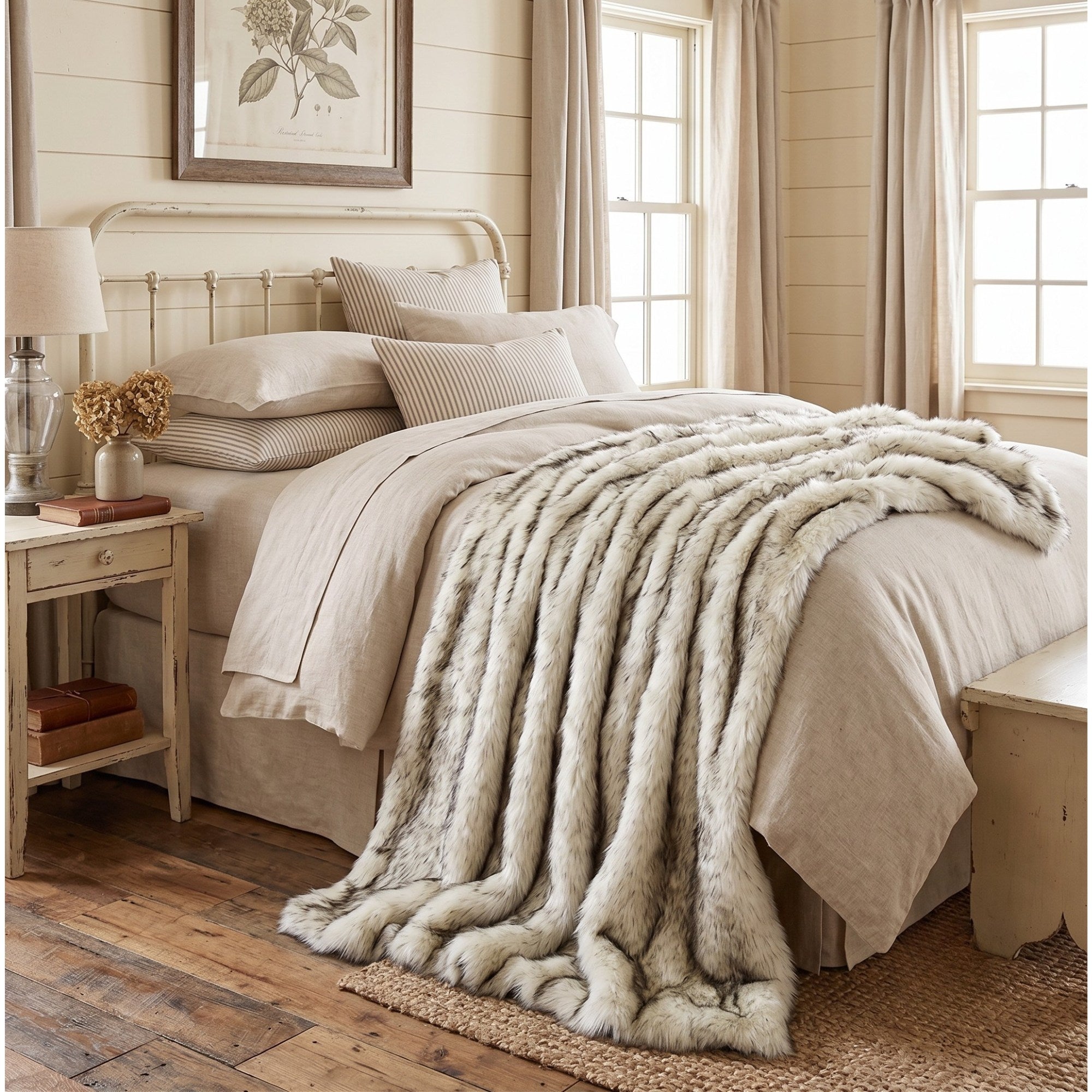 A plush faux fur throw blanket in a light beige-gray color is draped over a neatly made bed with neutral-toned linens in a rustic, warm bedroom setting.