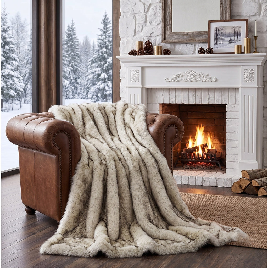 A plush, thick faux-fur throw blanket in a warm cream and beige tone is draped over a brown leather armchair in a cozy living room setting with a lit fireplace and snowy window view.