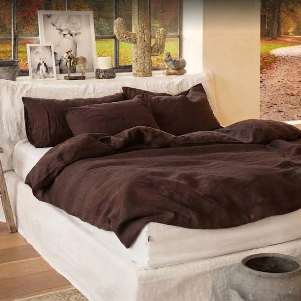 A bed dressed in dark brown plush bedding, including a duvet cover and matching pillows, styled in a rustic bedroom setting with a white bed skirt and window backdrop.