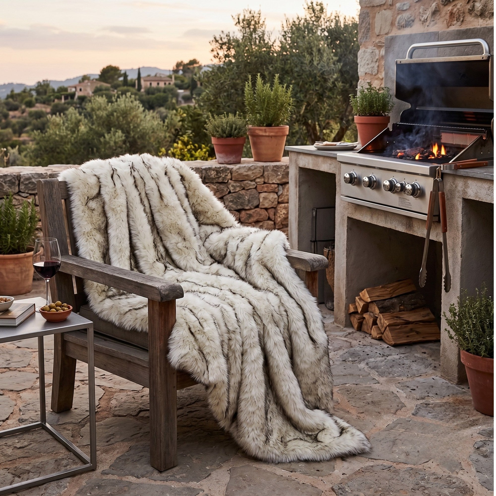 A plush faux-fur throw blanket in cream and taupe tones is draped over a rustic wooden outdoor chair on a stone patio. The setting includes a built-in grill, potted plants, and a scenic view in the background at sunset.