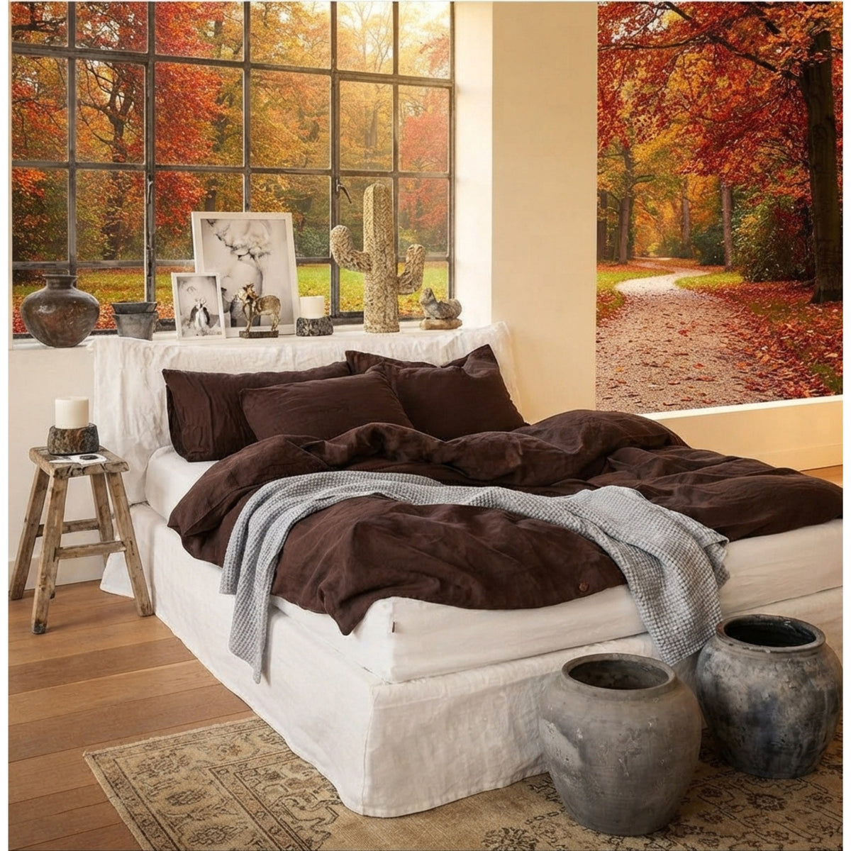 A styled bedroom scene featuring a bed dressed in dark brown bedding and a light gray knit throw, set against a wall mural of an autumn forest path. The room includes a large window with fall foliage outside, a rustic wooden stool, and decorative ceramic pots.