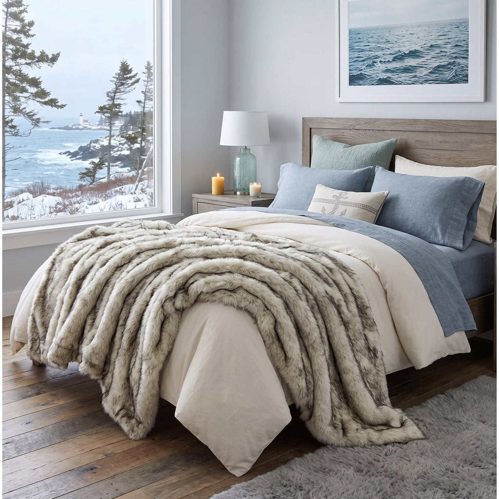 A styled bedroom scene showing a bed dressed in cream bedding with a plush faux-fur throw blanket draped across the foot, blue pillows, a wooden headboard, and a large window overlooking a snowy coastal landscape.