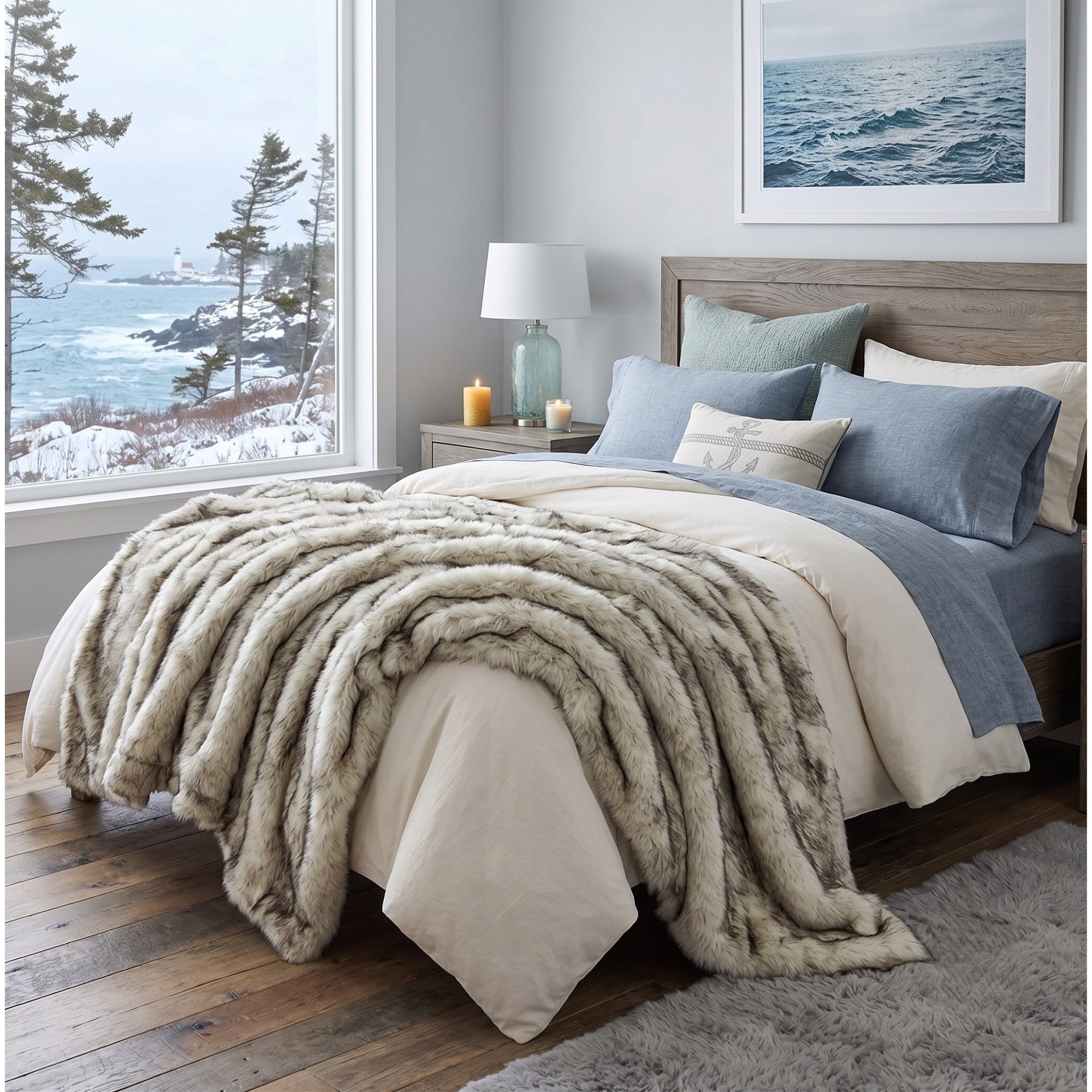 A styled bedroom scene showing a bed dressed in cream bedding with a plush faux-fur throw blanket draped across the foot, blue pillows, a wooden headboard, and a large window overlooking a snowy coastal landscape.