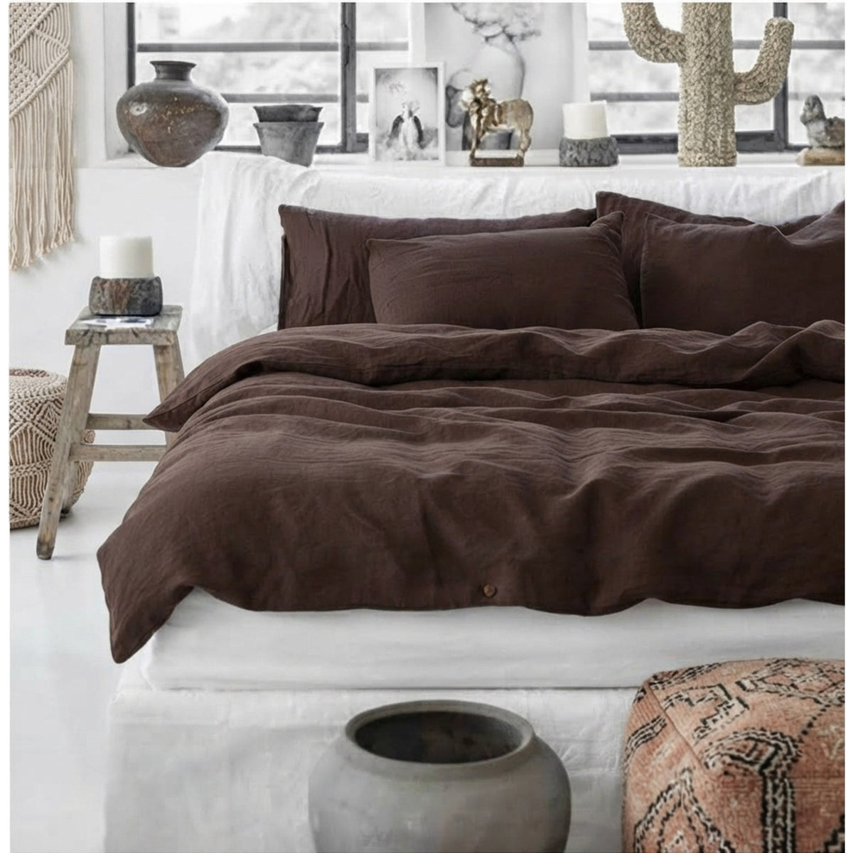 A bed dressed in dark brown bedding, including a duvet cover and matching pillowcases, styled in a bright, modern bedroom with white walls, decorative vases, and a small wooden stool.