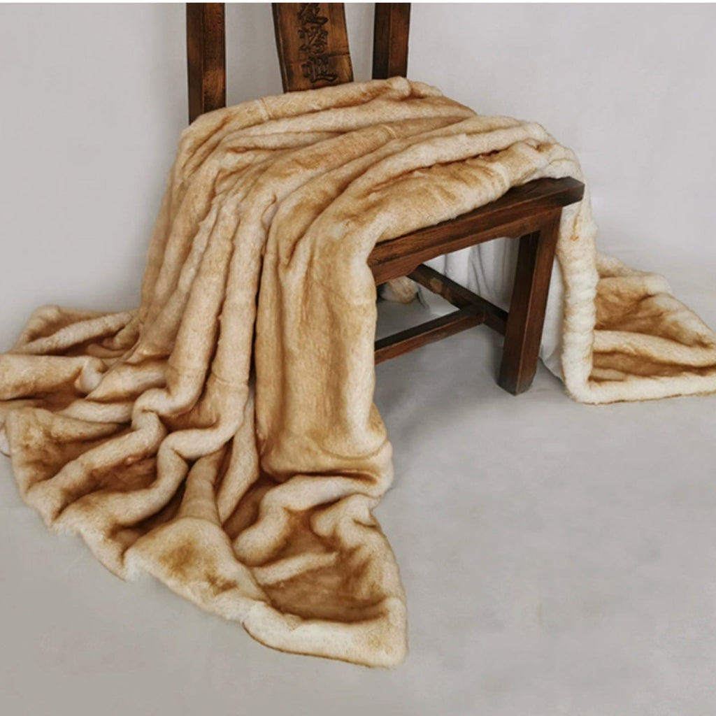 A plush faux-fur style throw blanket in warm beige and tan tones draped over a dark wooden chair against a plain light gray studio background.