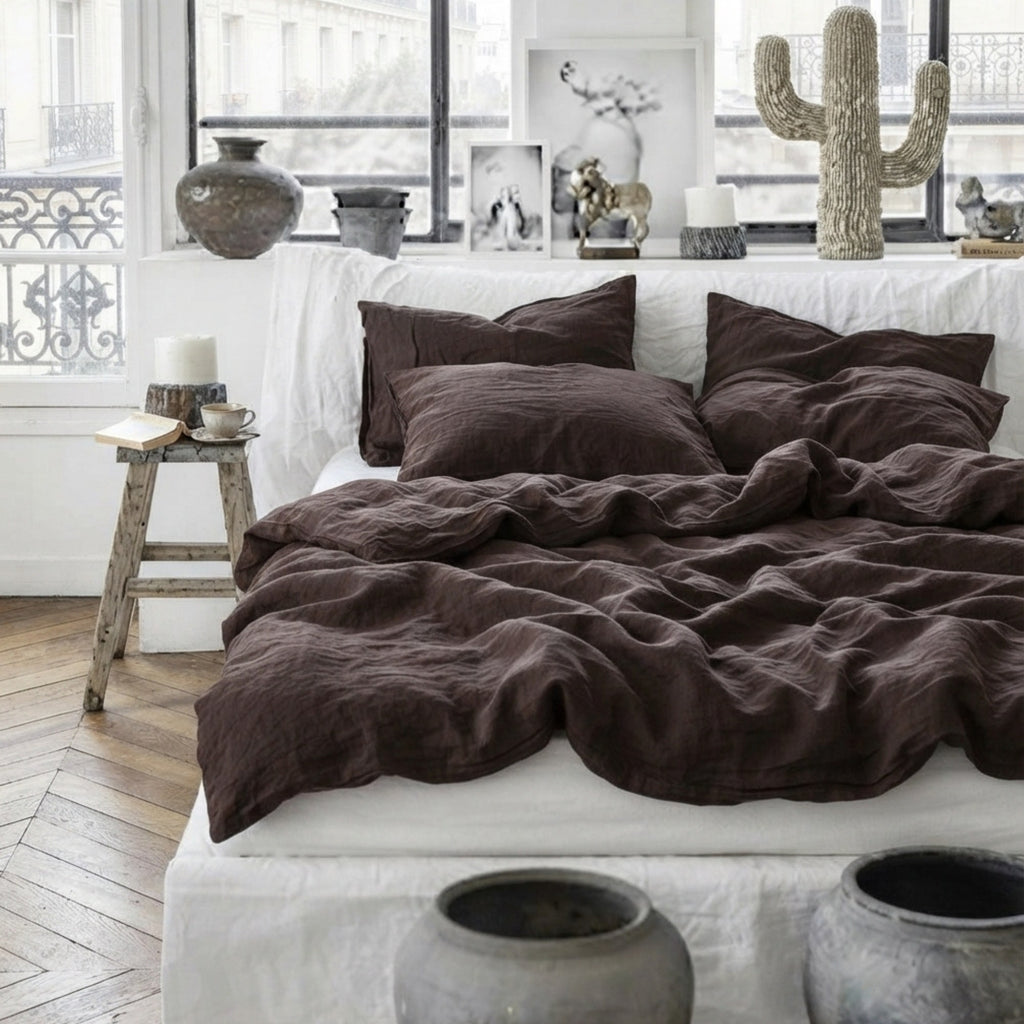 A bed dressed in dark brown crinkled linen bedding, including a duvet cover and matching pillowcases, styled in a bright, modern bedroom with white walls, large windows, and decorative vases.