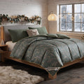A cozy bedroom scene showing a bed dressed in a green damask-patterned comforter with matching pillows, set in a rustic wood-paneled room with a window, floor lamp, and holiday garland.