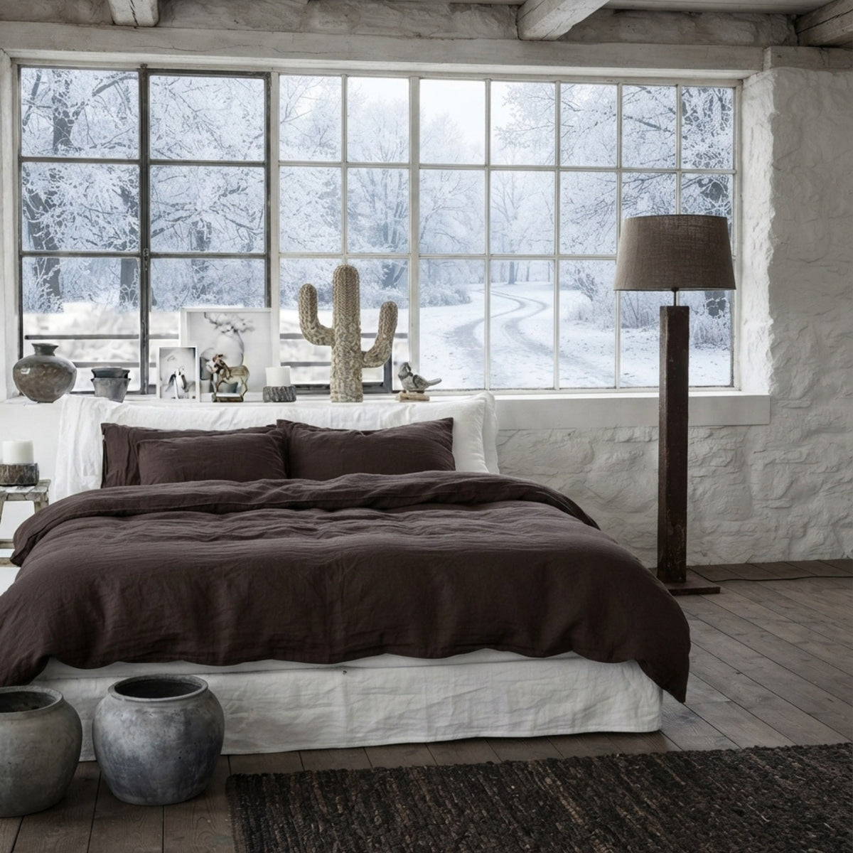 A bed dressed in dark brown and white bedding, styled in a rustic bedroom with a large window showing a snowy outdoor view, a floor lamp, and decorative ceramic pots.