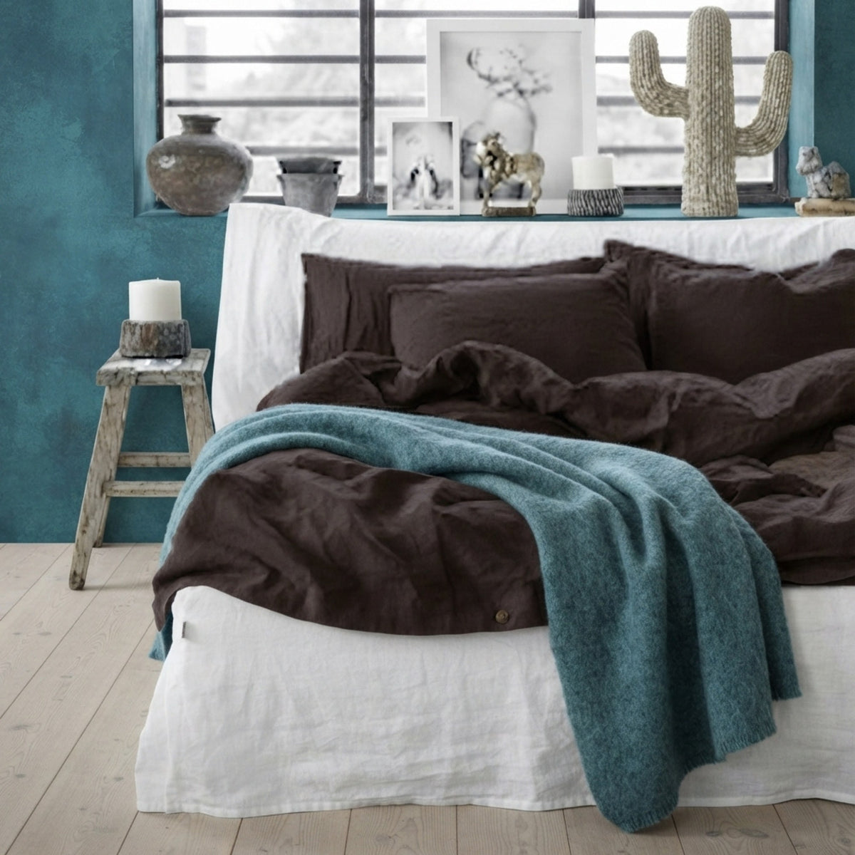 A styled bed in a bedroom setting with dark brown bedding, including pillows and a duvet, layered with a teal throw blanket. The room features a teal wall, a rustic wooden stool with a candle, and a window in the background.