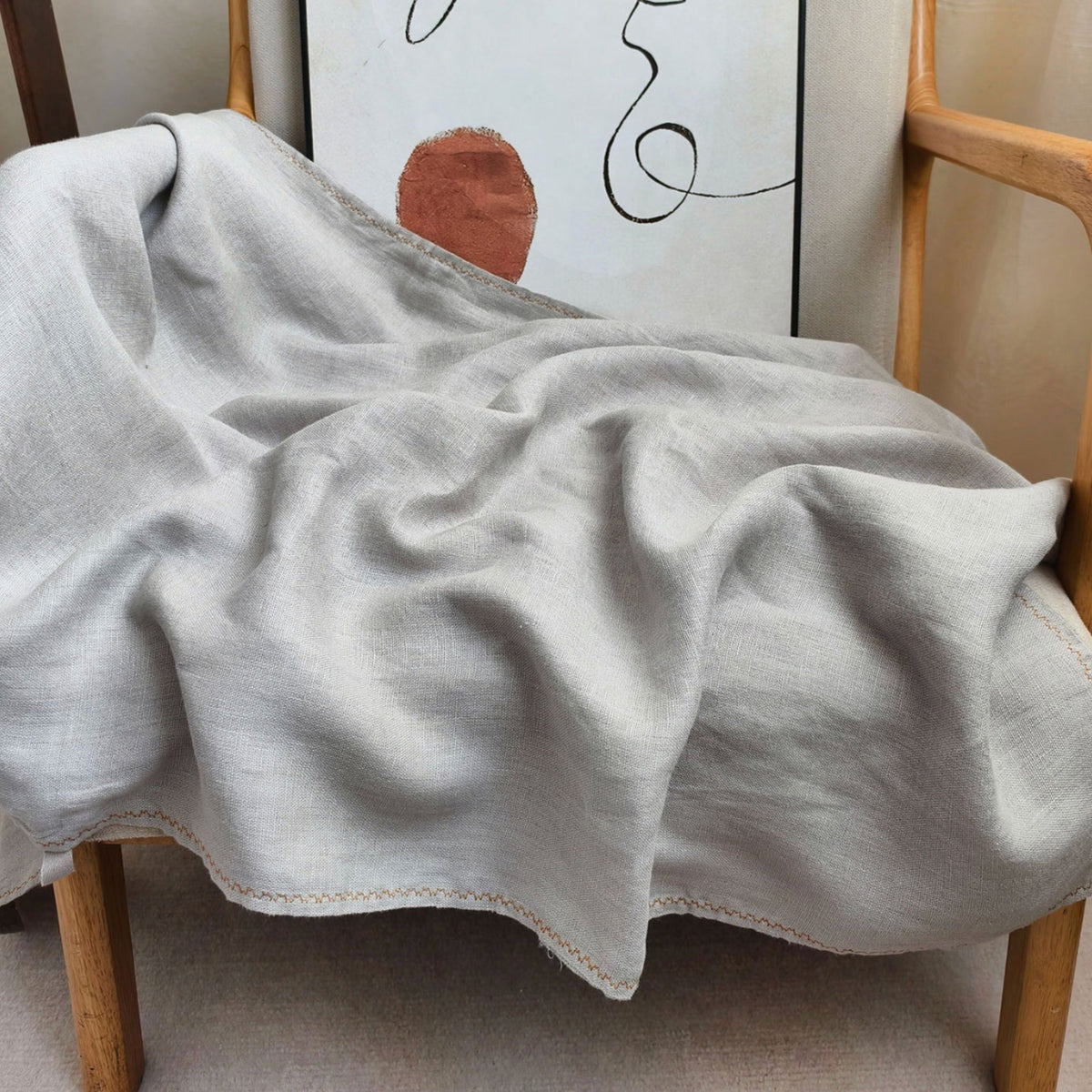 A light gray linen throw blanket with delicate hand-stitched edges draped over a wooden chair in a styled indoor setting.