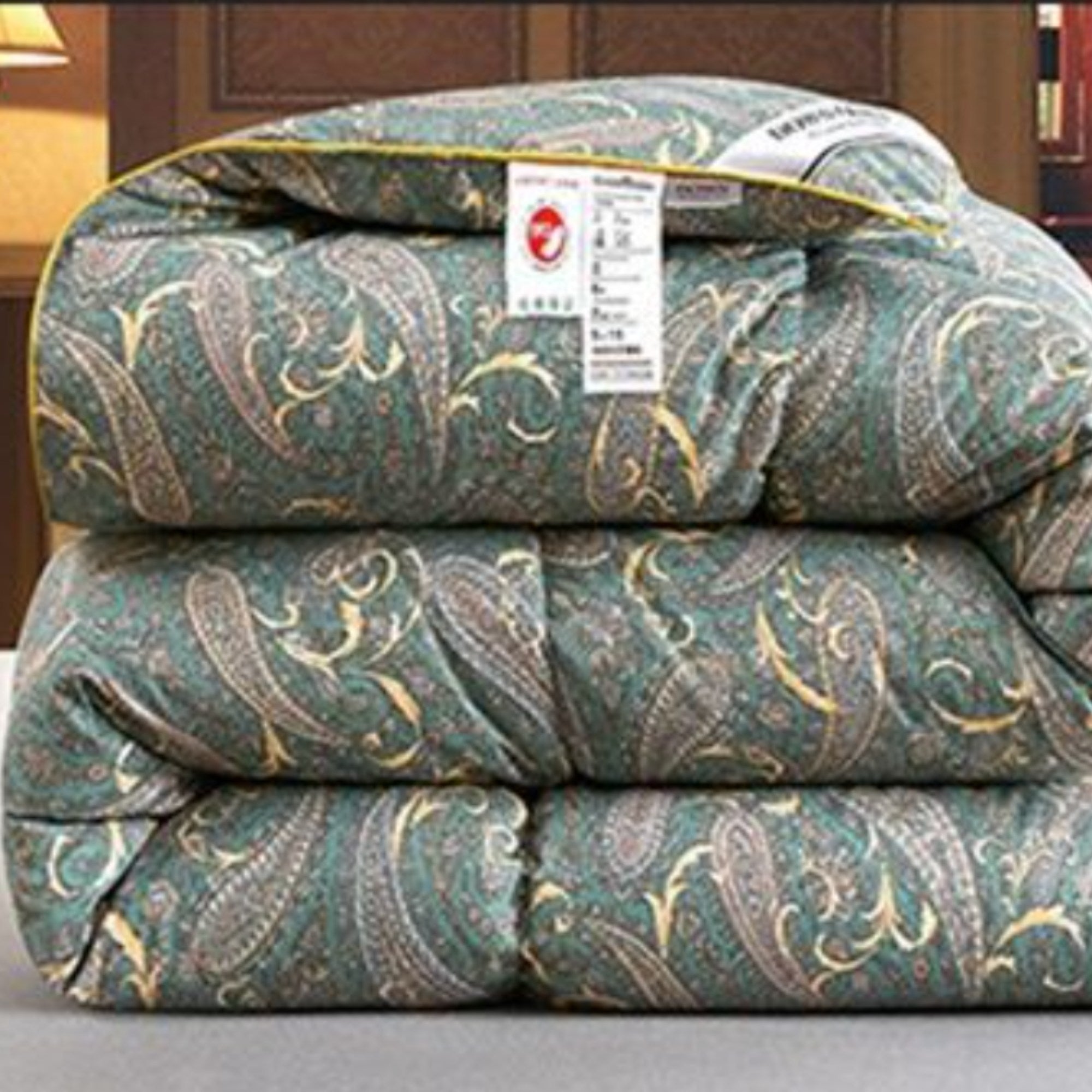 A folded quilted comforter with a teal green paisley pattern and gold trim, featuring a white product tag with a red logo.