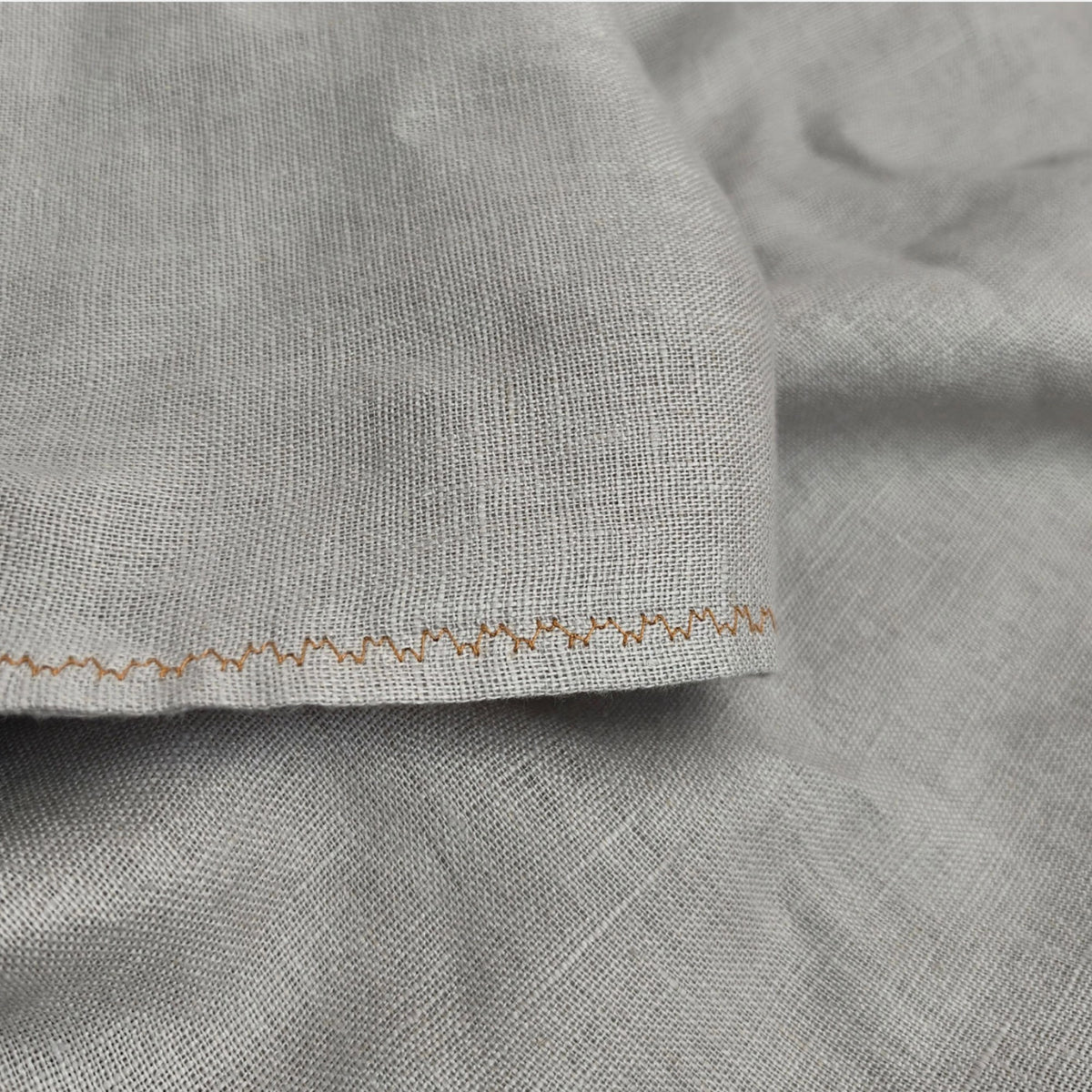 Close-up of light gray woven fabric with a visible zigzag stitched edge and subtle tan thread detail.
