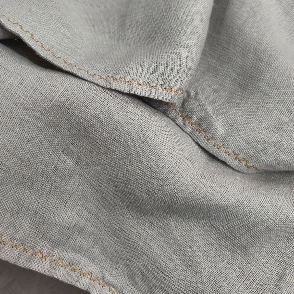 Close-up of a light gray linen fabric with visible texture and tan zigzag stitching along the edges.