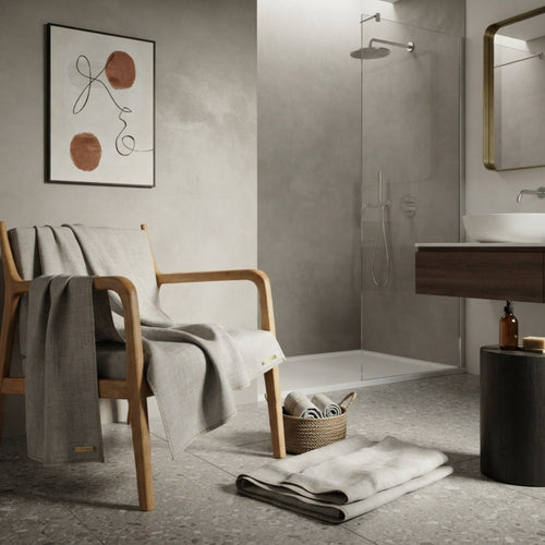 A modern bathroom interior with a light wood armchair draped in a gray linen throw, a folded matching towel on the floor, a small woven basket, a wall-mounted framed abstract print, and a glass shower enclosure in the background.