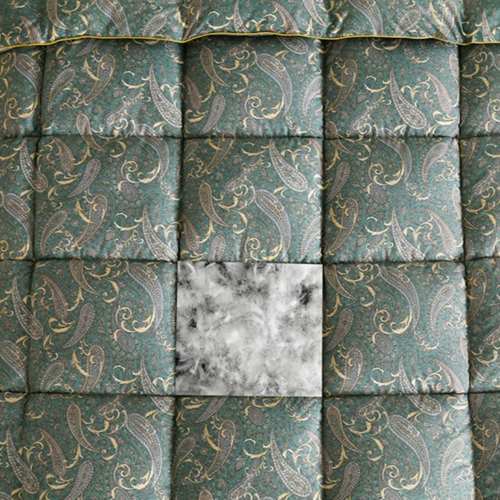 Close-up of a quilted fabric surface with a teal-green background and an allover paisley pattern in muted gold and gray. A square patch of white-and-gray marbled material is centered over the patterned fabric.
