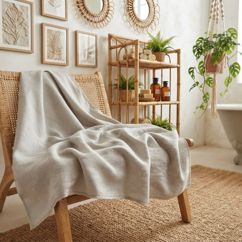 A light beige linen throw blanket is draped over a woven rattan armchair in a bright, boho-style room with a wooden shelf, plants, and framed botanical prints.