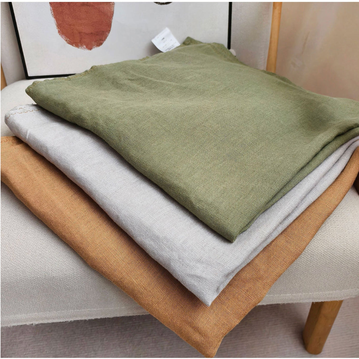 Three folded linen fabric pieces in olive green, light gray, and tan are stacked on a light-colored upholstered chair.