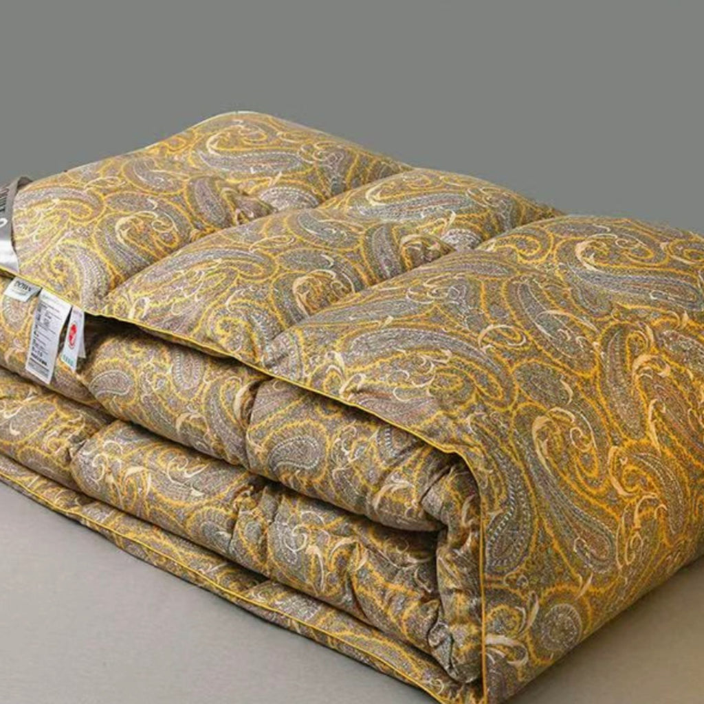 A folded quilted comforter or bedspread with a yellow-gold and gray paisley pattern, shown on a plain gray studio background. A paper tag is attached to the left side.