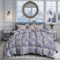 A bed dressed in a blue and beige paisley-patterned comforter set with matching pillows, styled in a bright bedroom with light wood walls, bedside tables, wall sconces, and large windows overlooking a snowy mountain and ocean view.
