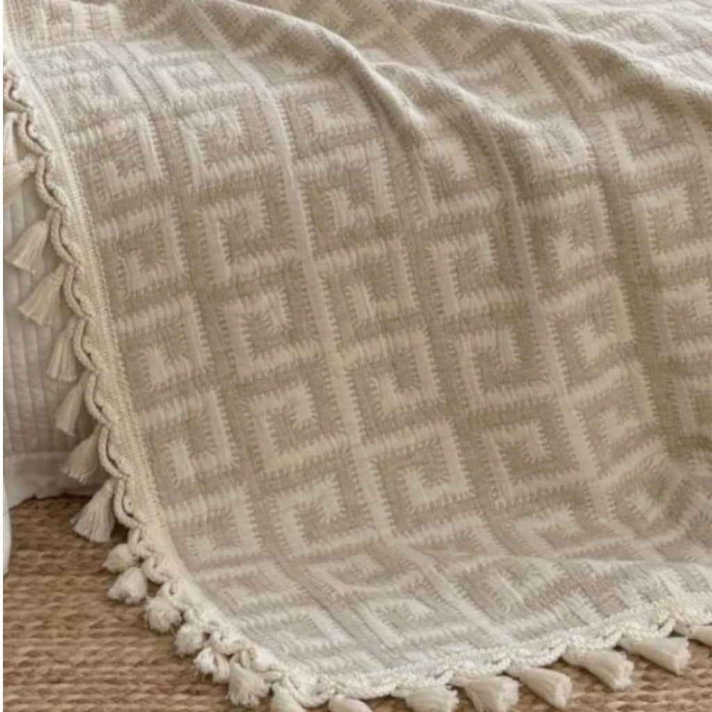 A close-up of a cream and beige woven throw blanket with a geometric pattern and tasseled edge, draped over a bed on a woven rug.