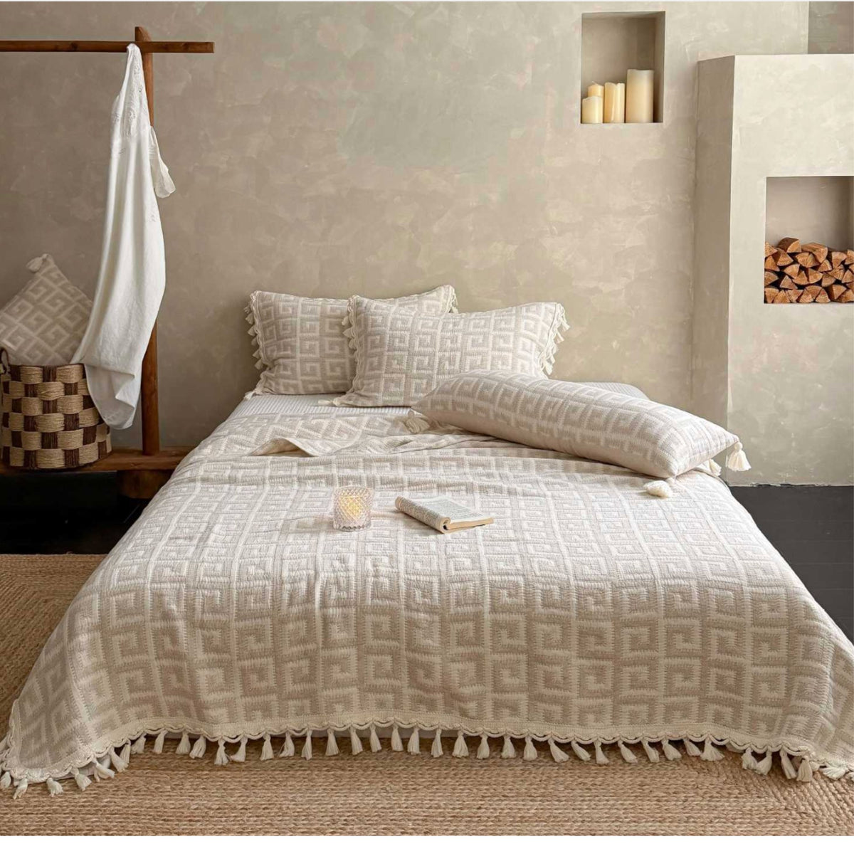 A neatly made bed in a styled bedroom setting, covered with a cream and beige patterned quilt and matching pillowcases. The room features textured plaster walls, a wooden hanger with a white shirt, a woven basket, and a built-in fireplace niche with logs.