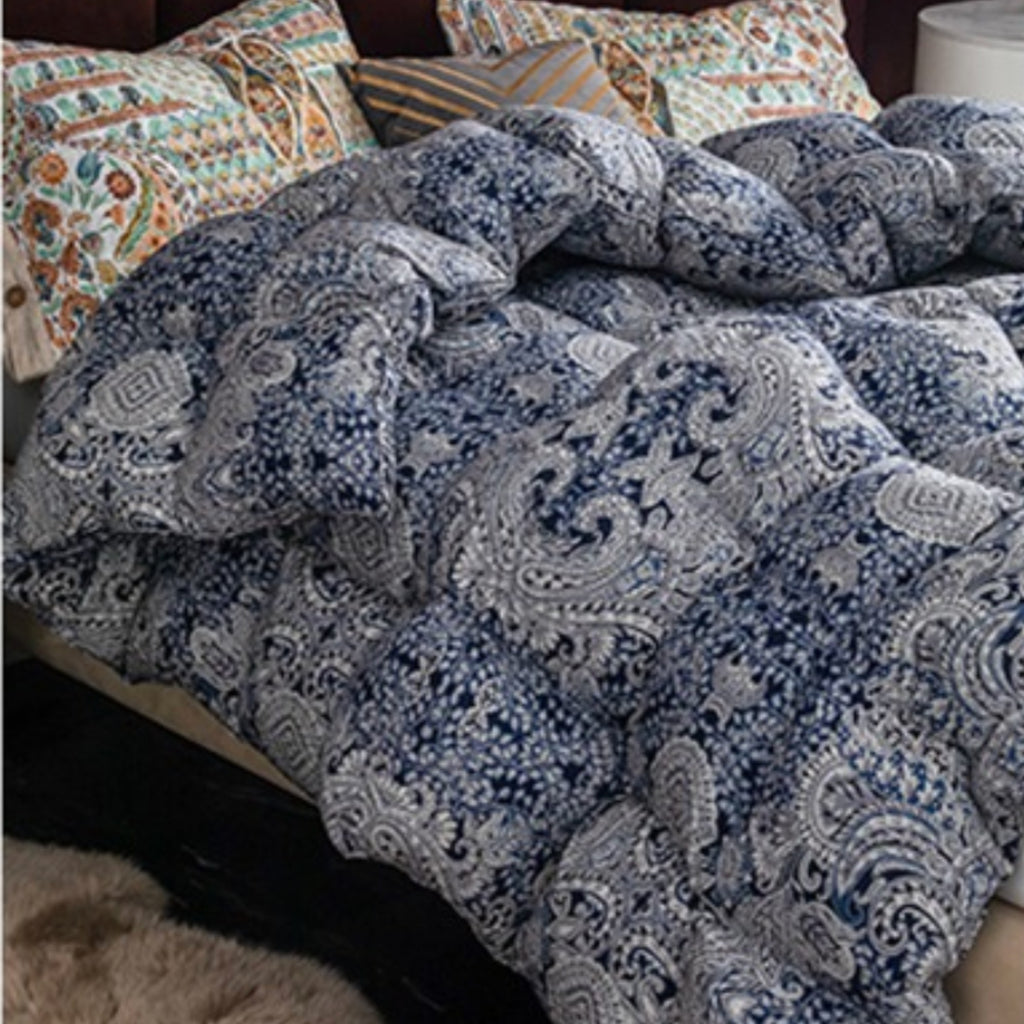 A close-up of a bed with a blue and white paisley patterned comforter or duvet, styled with patterned pillows and a dark headboard in a bedroom setting.