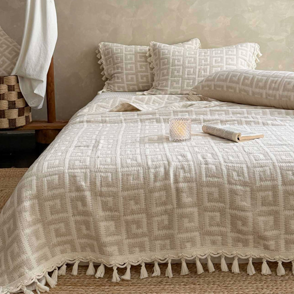A cream-colored textured blanket with a geometric pattern and tassel fringe is styled on a bed in a warm, rustic bedroom setting. Matching patterned pillows and a pillowcase are arranged behind it, with a small book and a lit candle on the blanket.