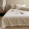 A cream-colored textured blanket with a geometric pattern and tassel fringe is styled on a bed in a warm, rustic bedroom setting. Matching patterned pillows and a pillowcase are arranged behind it, with a small book and a lit candle on the blanket.