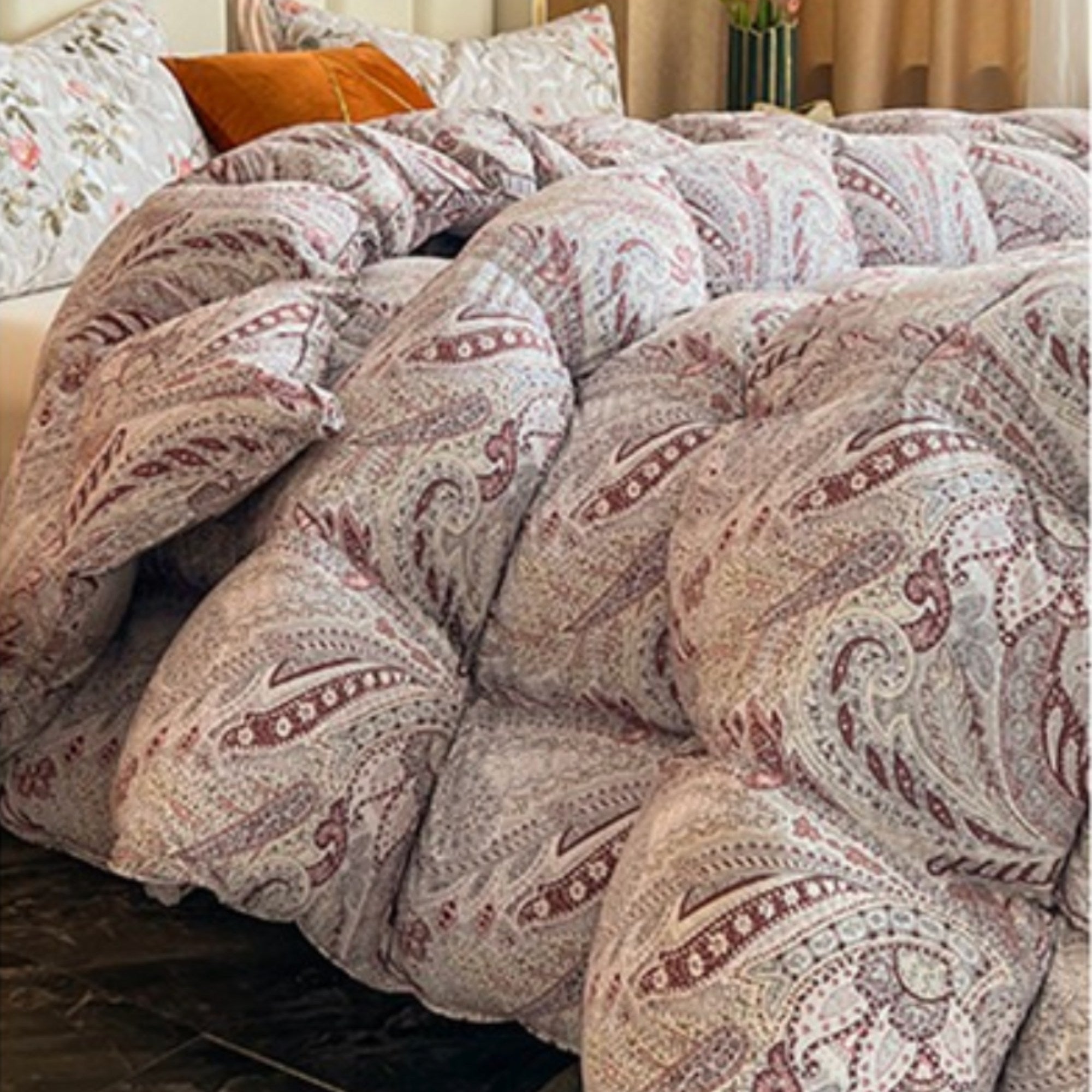 A close-up of a fluffy quilted comforter with a paisley pattern in muted dusty pink, beige, and gray tones, styled on a bed with pillows in a softly lit bedroom.